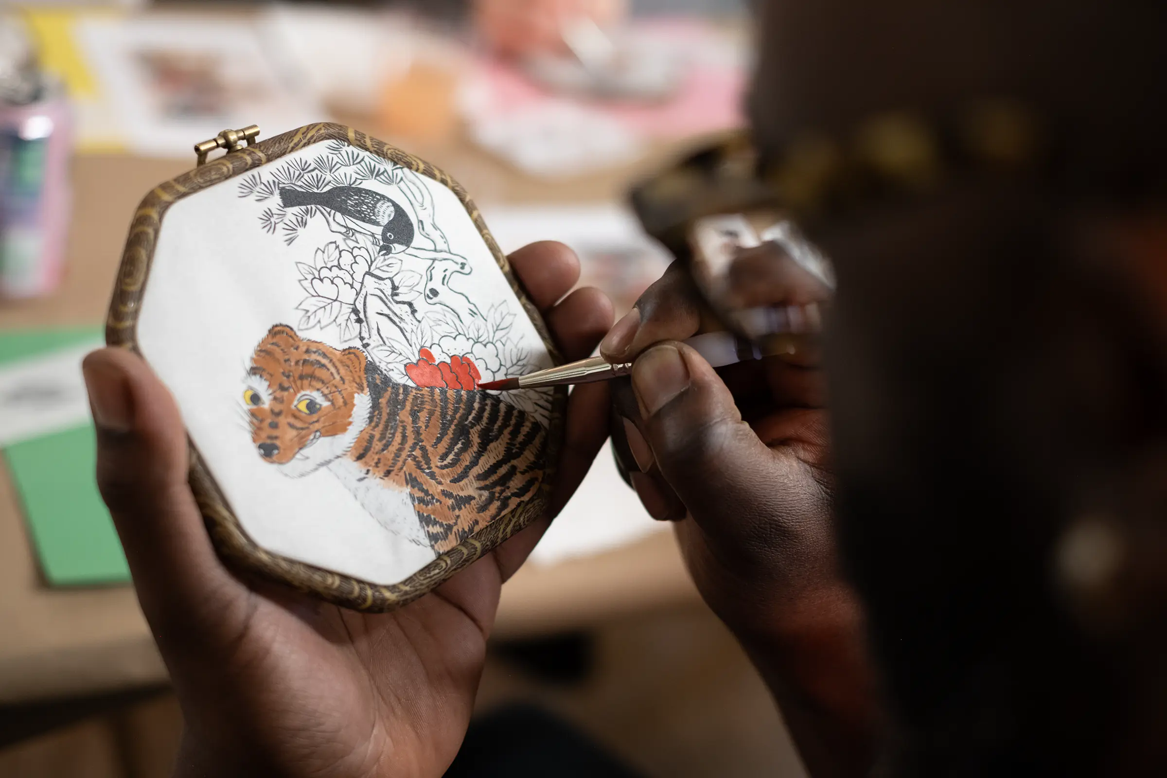 A man painting a tiger, magpie, and peonies in the traditional Korean painting style on traditional paper, set in an ornate wooden frame.