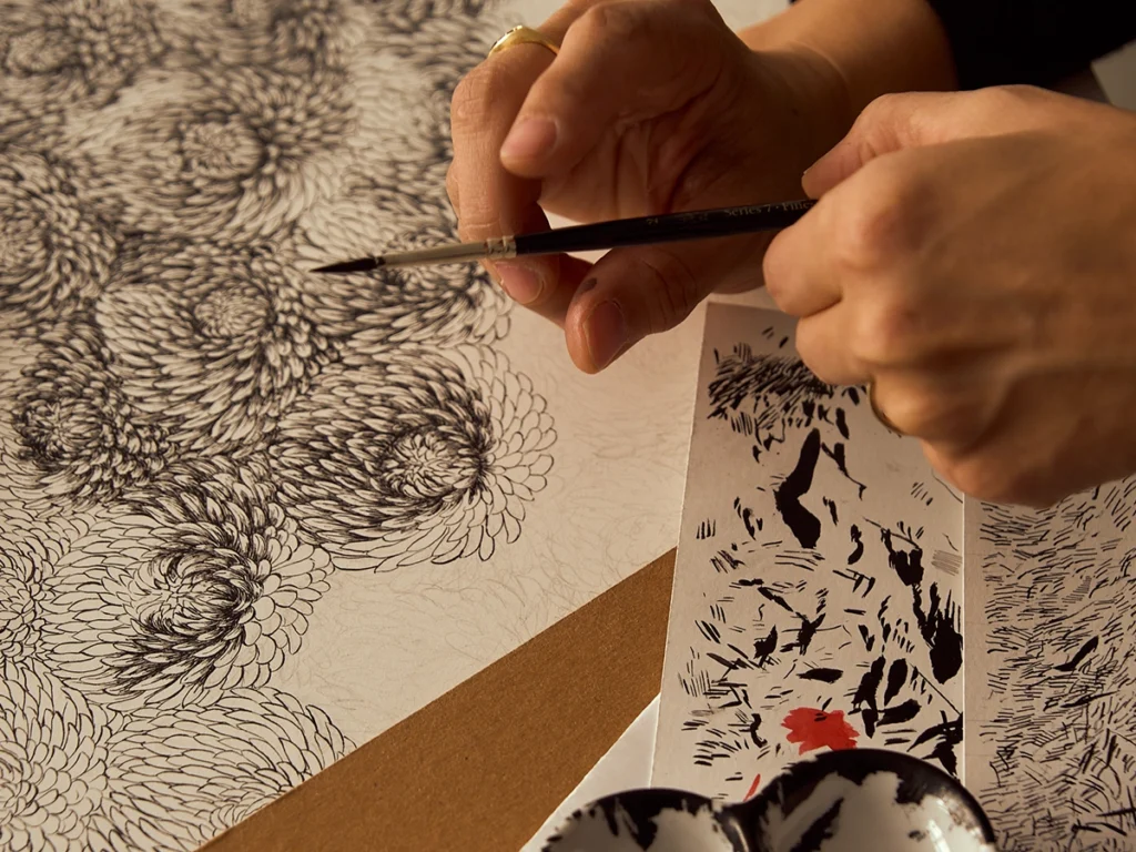 Author holding a brush with both hands above a drawing and ink on a table.