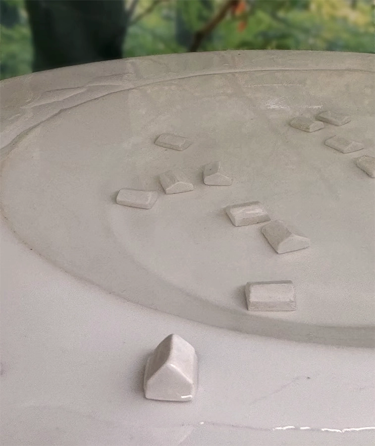 Ceramic installation with miniature house blocks submerged in a sunken circular space within a raised form.