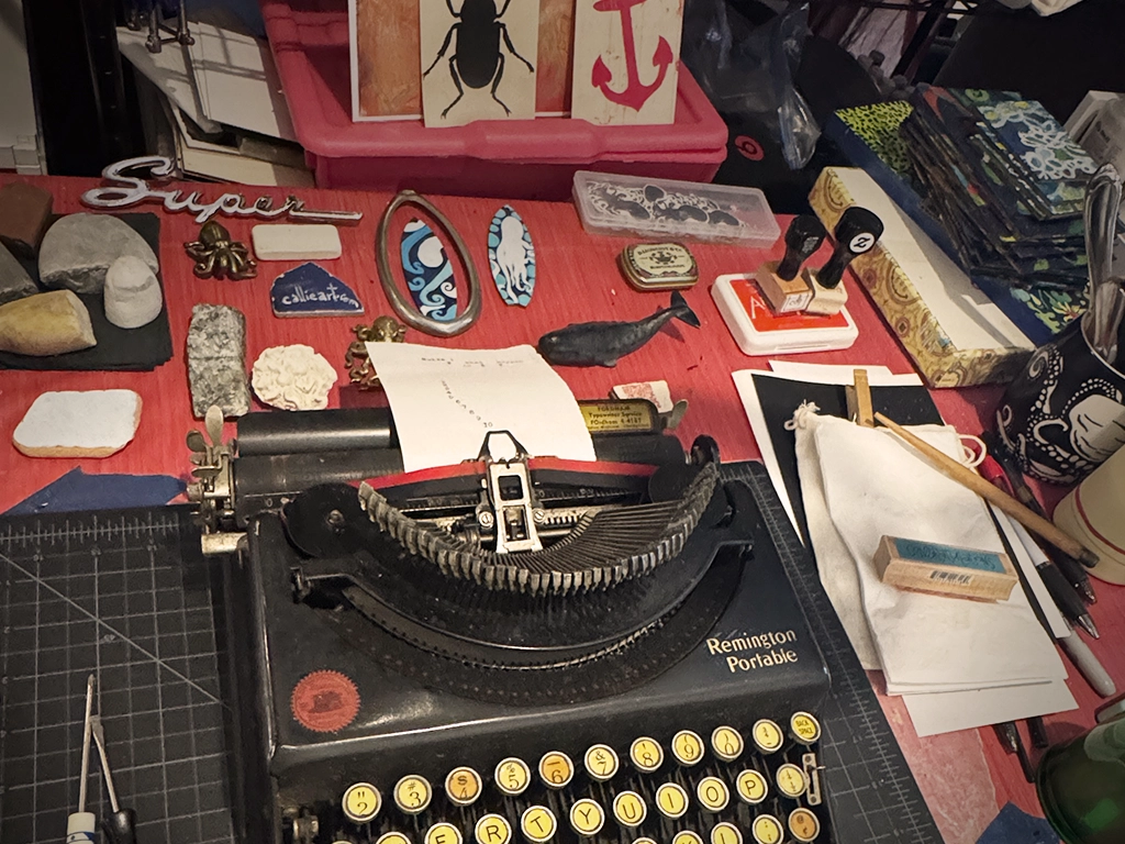Artist’s worktable with a vintage typewriter, stamps, tools, sketches, and collected objects.