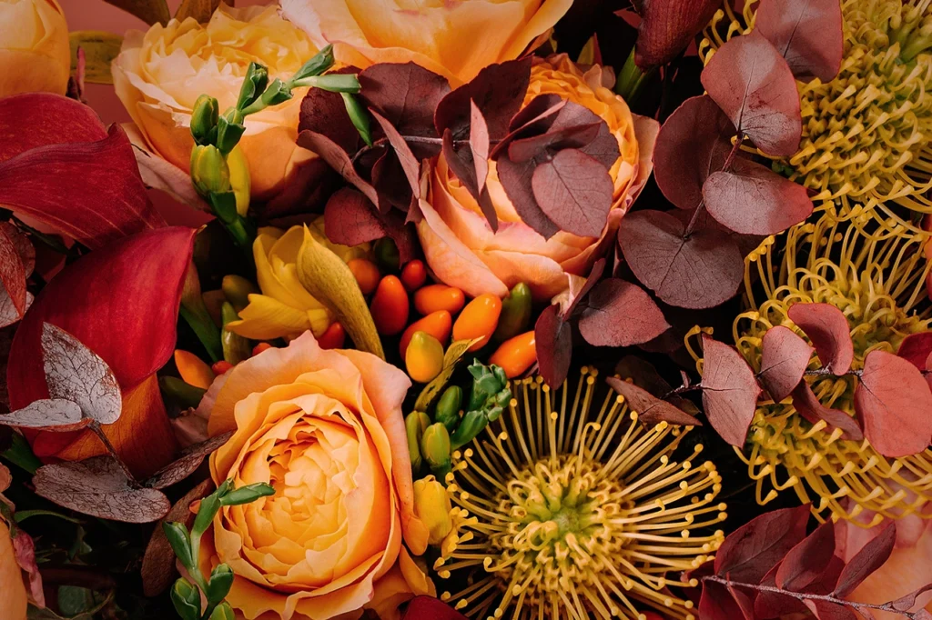 Close-up of a fall floral arrangement with peach roses, burgundy leaves, orange berries, and textured seasonal blooms.