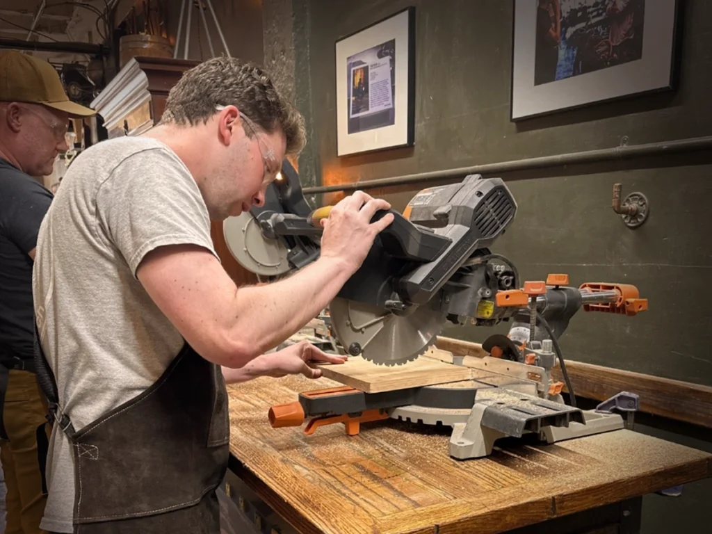 A participant cutting a piece of oak with a miter saw during a hands-on woodworking class.